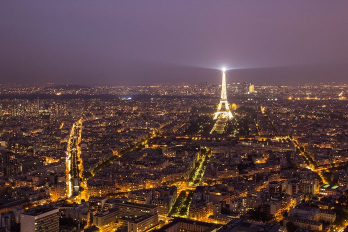 Une vue aérienne nocturne de Paris montre la ville illuminée par les lampadaires. La Tour Eiffel se dresse au centre de la scène, illuminée par les projecteurs, tandis que des monuments tels que l'Hôtel Renoir Paris se fondent dans les avenues animées qui s'étendent vers l'horizon brumeux. 