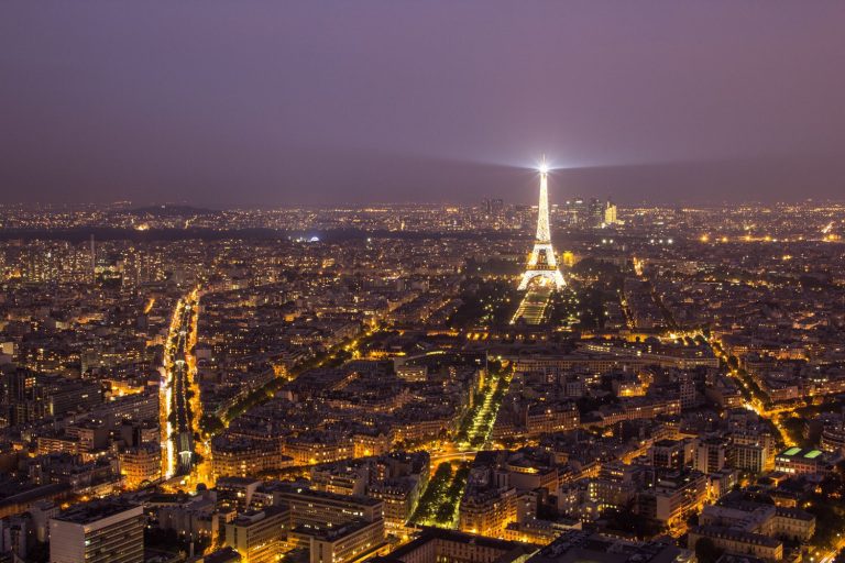 Une vue aérienne nocturne de Paris montre la ville illuminée par les lampadaires. La Tour Eiffel se dresse au centre de la scène, illuminée par les projecteurs, tandis que des monuments tels que l'Hôtel Renoir Paris se fondent dans les avenues animées qui s'étendent vers l'horizon brumeux. 