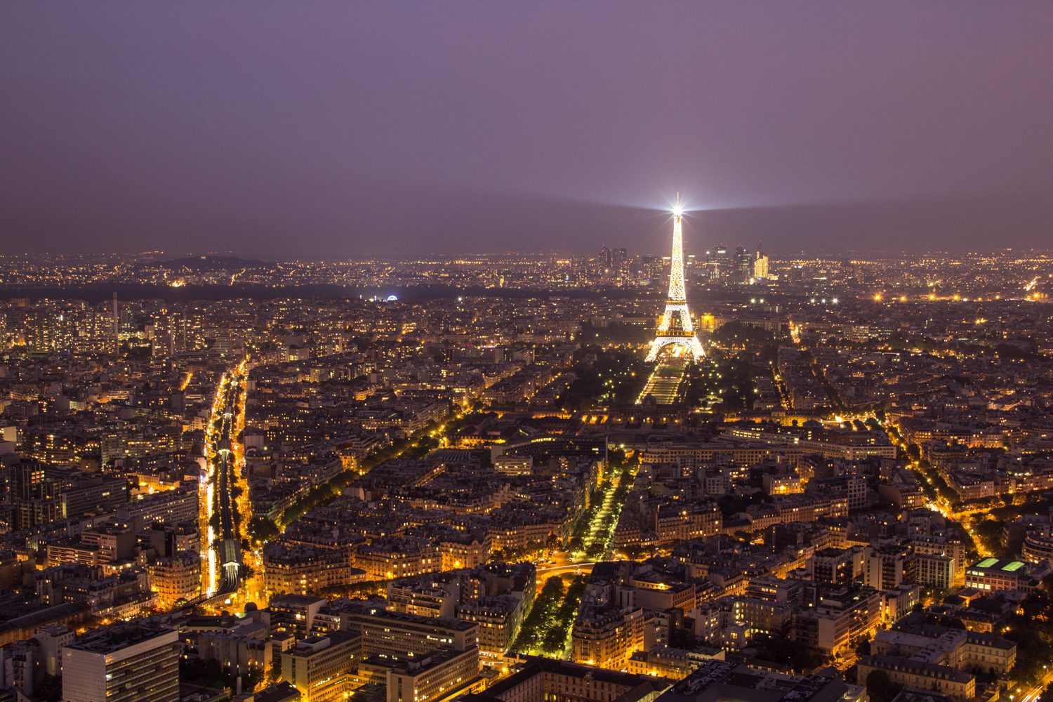 IMG_9008-1-6 Une vue aérienne nocturne de Paris montre la ville illuminée par les lampadaires. La Tour Eiffel se dresse au centre de la scène, illuminée par les projecteurs, tandis que des monuments tels que l'Hôtel Renoir Paris se fondent dans les avenues animées qui s'étendent vers l'horizon brumeux.