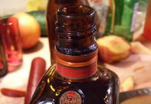 Cointreau, la “marque mondiale” A close-up of a brown glass Cointreau liqueur bottle, the iconic marque mondiale, sits on a wooden counter. Its label and orange cap stand out amid blurred cocktail-making tools like fruits, a red muddler, and a knife.