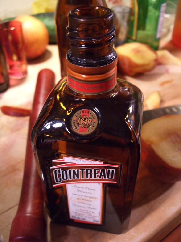 Gros plan sur une bouteille de liqueur Cointreau en verre brun, la marque mondiale emblématique, posée sur un comptoir en bois. Son étiquette et son bouchon orange se détachent au milieu d'outils de préparation de cocktails flous tels que des fruits, un mélangeur rouge et un couteau. 