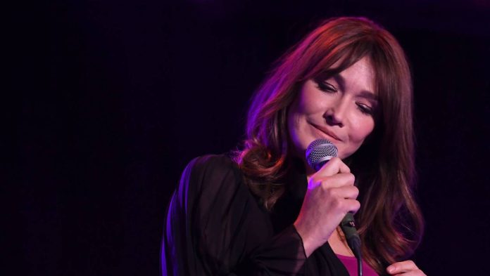 Une femme aux longs cheveux bruns chante sur scène, canalisant la poésie de Carla Bruni. Tenant un micro à proximité, elle porte un haut transparent sombre sur une chemise rose, les yeux fermés, tandis que des lumières violettes créent une atmosphère douce et intime. 