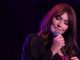 Carla Bruni, version «poetry» A woman with long brown hair sings on stage, channeling the poetry of Carla Bruni. Holding a microphone close, she wears a dark sheer top over a pink shirt, her eyes closed as purple lights create a soft and intimate atmosphere.