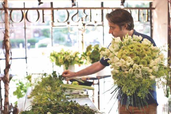 Un homme arrange des fleurs dans une boutique lumineuse et rustique inspirée par Christian Tortu. Il tient un grand bouquet rond de lys blancs et de feuillage vert, tandis que d'autres fleurs sont étalées sur une table en bois et que la lumière du soleil traverse les fenêtres. 