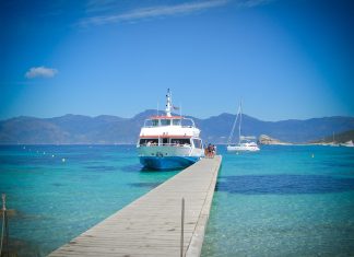 Corsica Travel Guide: Hiking the GR20 & The Island of Beauty A white and blue ferry is docked at the end of a wooden pier over clear turquoise water, with people boarding. In the background are mountains under a bright blue sky and another boat anchored further out. The scene is calm and sunlit.