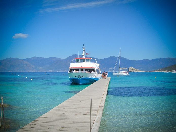 Un ferry blanc et bleu est amarré au bout d'une jetée en bois sur une eau claire et turquoise, et des personnes montent à bord. À l'arrière-plan, on aperçoit des montagnes sous un ciel d'un bleu éclatant et un autre bateau ancré plus loin. La scène est calme et ensoleillée.  