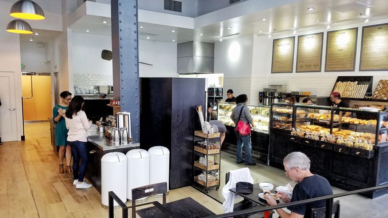 Un café-boulangerie moderne inspiré par Pascal Rigo, le célèbre boulanger de San Francisco, avec des clients qui commandent à un comptoir de pâtisseries. Le personnel travaille derrière des vitrines, tandis qu'un homme est assis devant. L'espace présente des planchers en bois clair, des murs blancs et des accents industriels.  
