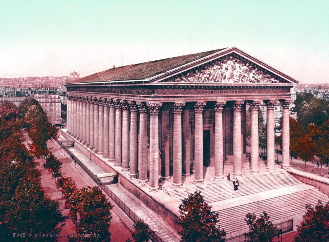 madeleine-de-napoleon A neoclassical building with tall Corinthian columns, broad steps, and a sculpted pediment. Trees line the walkway as people stand near the entrance, while the Parisian cityscape—echoing the gloire of Napoléon 1er—spreads under a pale sky.