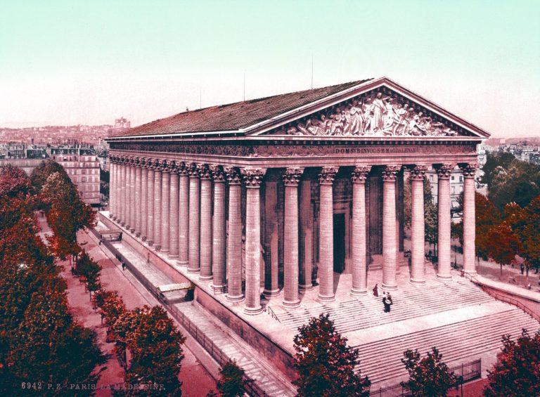Un bâtiment néoclassique avec de hautes colonnes corinthiennes, de larges marches et un fronton sculpté. Des arbres bordent l'allée tandis que les gens se tiennent près de l'entrée, tandis que le paysage urbain parisien - qui fait écho à la gloire de Napoléon 1er - s'étend sous un ciel pâle. 