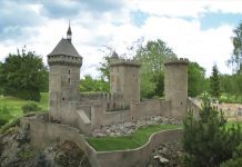 Comment visiter la France en trois heures Un château médiéval en pierre avec trois tours rondes et une flèche à toit pointu se dresse sur une colline rocheuse, idéal pour ceux qui souhaitent visiter la France en trois heures lors d'un voyage express en France. Entouré d'arbres verts et d'herbe, ses créneaux et ses petites fenêtres ajoutent à son charme.