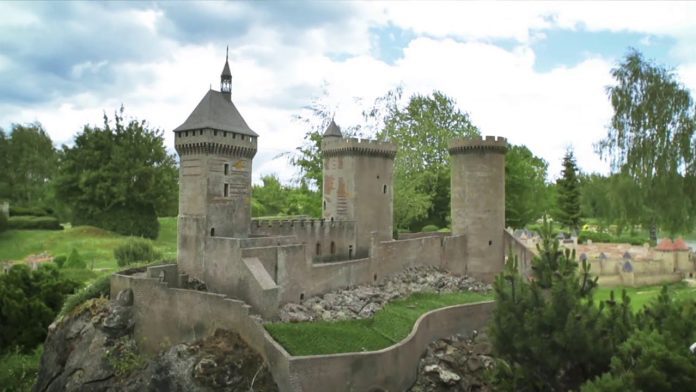 Un château médiéval en pierre avec trois tours rondes et une flèche à toit pointu se dresse sur une colline rocheuse, idéal pour ceux qui souhaitent visiter la France en trois heures lors d'un voyage express en France. Entouré d'arbres verts et d'herbe, ses créneaux et ses petites fenêtres ajoutent à son charme. 