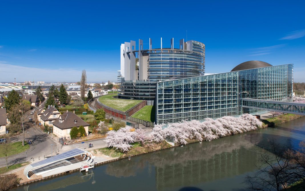 A modern glass and steel building with curved and rectangular sections sits beside a canal in Europe, lined with blooming cherry trees under a clear blue sky; small houses and a boat dock are visible nearby.