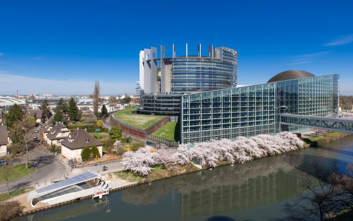 A modern glass and steel building with curved and rectangular sections sits beside a canal in Europe, lined with blooming cherry trees under a clear blue sky; small houses and a boat dock are visible nearby.