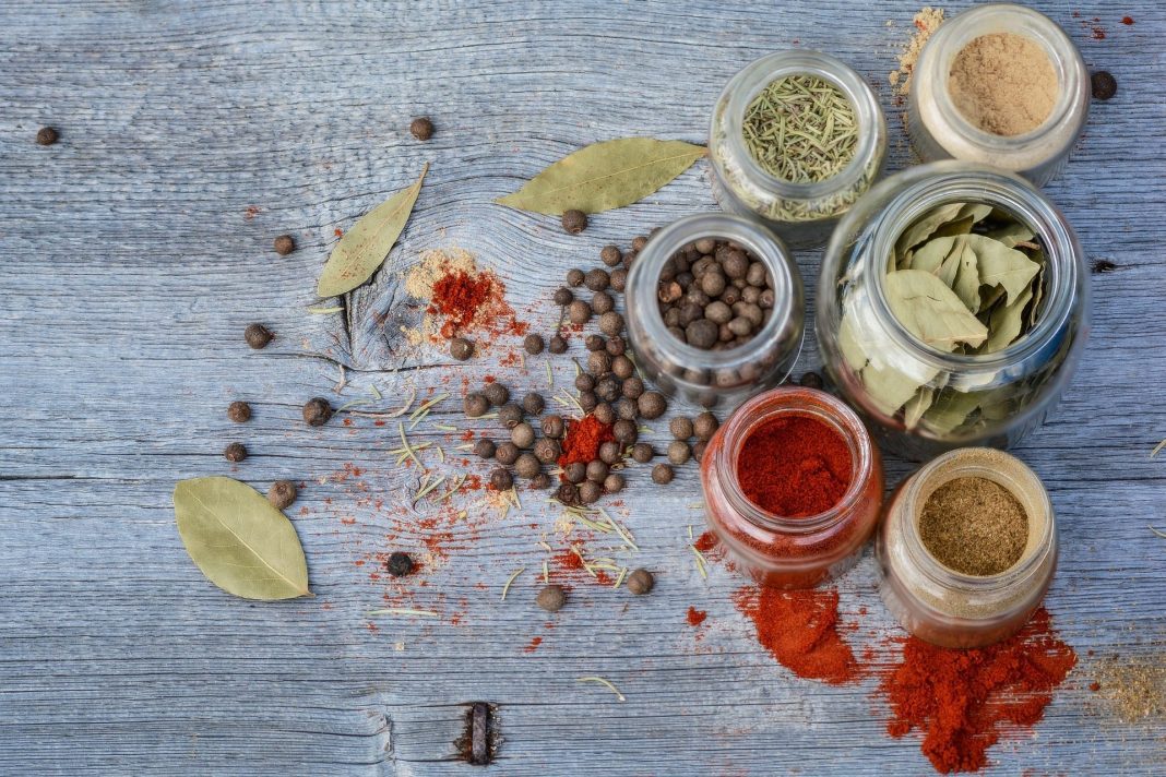 Glass jars filled with dried herbs and spices, including bay leaves, paprika, and peppercorns, are arranged on a rustic blue wooden surface—a scene perfect for any gastronomie lexique or traduction anglais français reference guide.