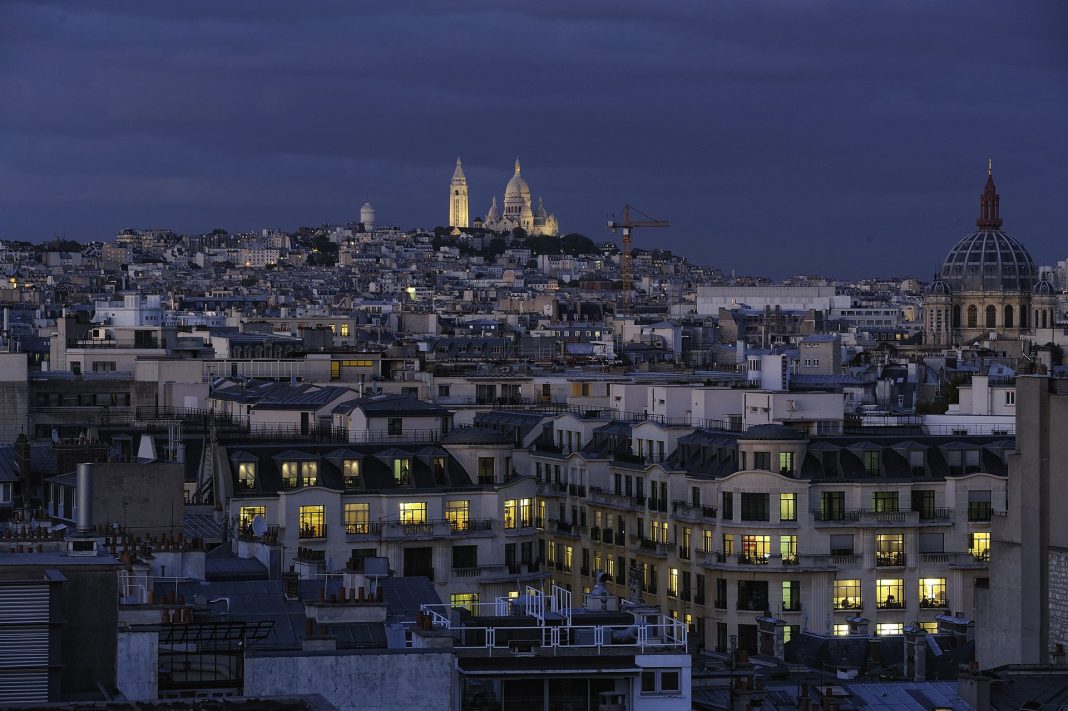 Un paysage urbain crépusculaire de Paris montre des rangées d'immeubles aux fenêtres lumineuses. La basilique du Sacré-Cœur, illuminée, se trouve au sommet de la colline de Montmartre, tandis qu'un aperçu des Champs-Élysées et une grue de construction ajoutent à la scène vibrante sous un ciel nuageux. 