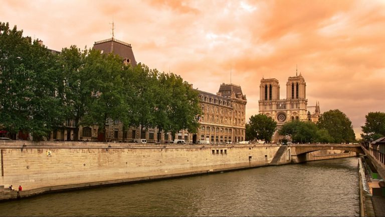 Vue de la Seine à Paris au coucher du soleil, avec des arbres feuillus et des bâtiments historiques en pierre près de l'hôtel Le Clos Medicis sur la rive gauche. La cathédrale Notre-Dame se dresse bien en évidence sous un ciel orange spectaculaire. 