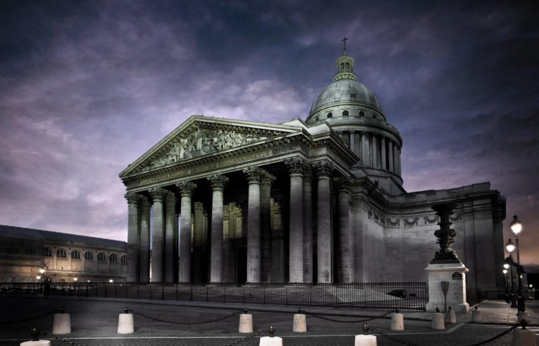 Un grand bâtiment néoclassique avec de hautes colonnes corinthiennes et un grand dôme central se dresse sous un ciel nuageux près de l'Hôtel Moderne Saint-Germain, sa façade éclairée donnant sur les rues pavées du cœur de Paris.