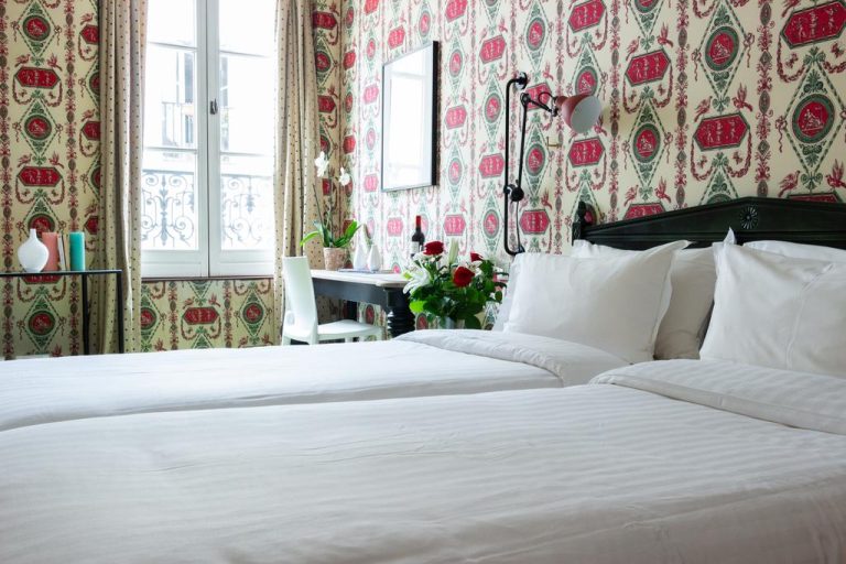 Une chambre confortable de l'hôtel Prince de Condé à Paris comprend deux lits jumeaux bien faits, du linge blanc et du papier peint à motifs rouges et verts. La lumière naturelle pénètre par un bureau sombre orné de fleurs, près d'une fenêtre aux rideaux beiges. 