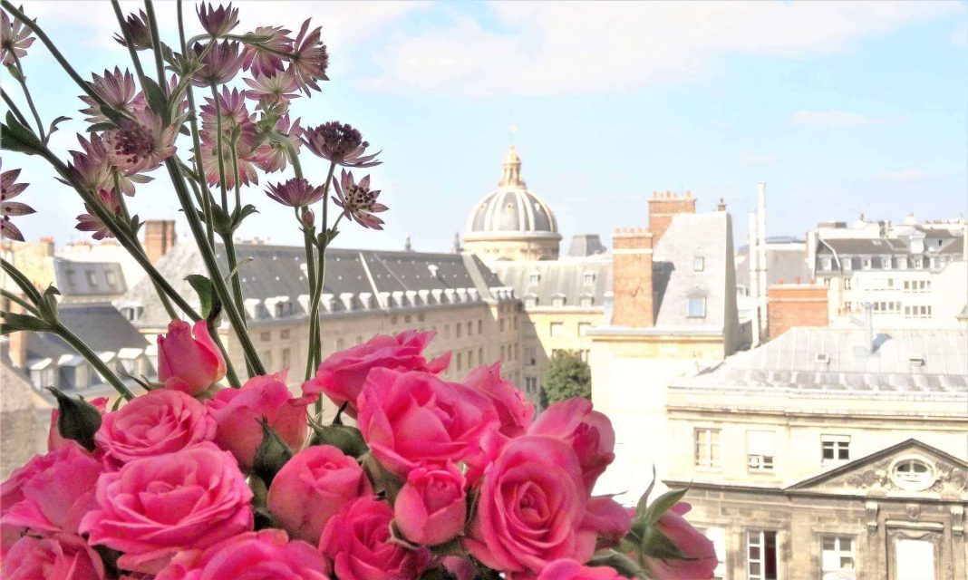 Un bouquet de roses roses éclatantes et de fleurs violettes se trouve au premier plan, entouré d'immeubles parisiens classiques aux toits gris. Le dôme de l'hôtel Prince de Conti s'élève à l'arrière-plan sous un ciel lumineux et partiellement nuageux. 