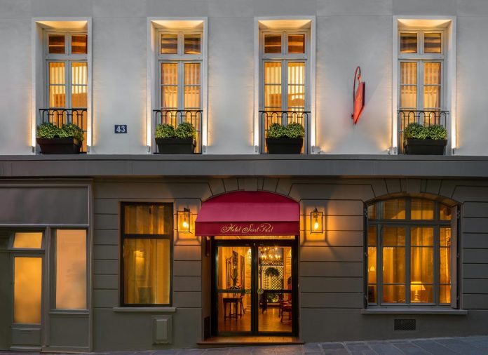 La façade de l'hôtel, chaudement éclairée la nuit, présente de hautes fenêtres avec des balustrades noires et des bacs à fleurs, un auvent rouge au-dessus de l'entrée sur lequel on peut lire 