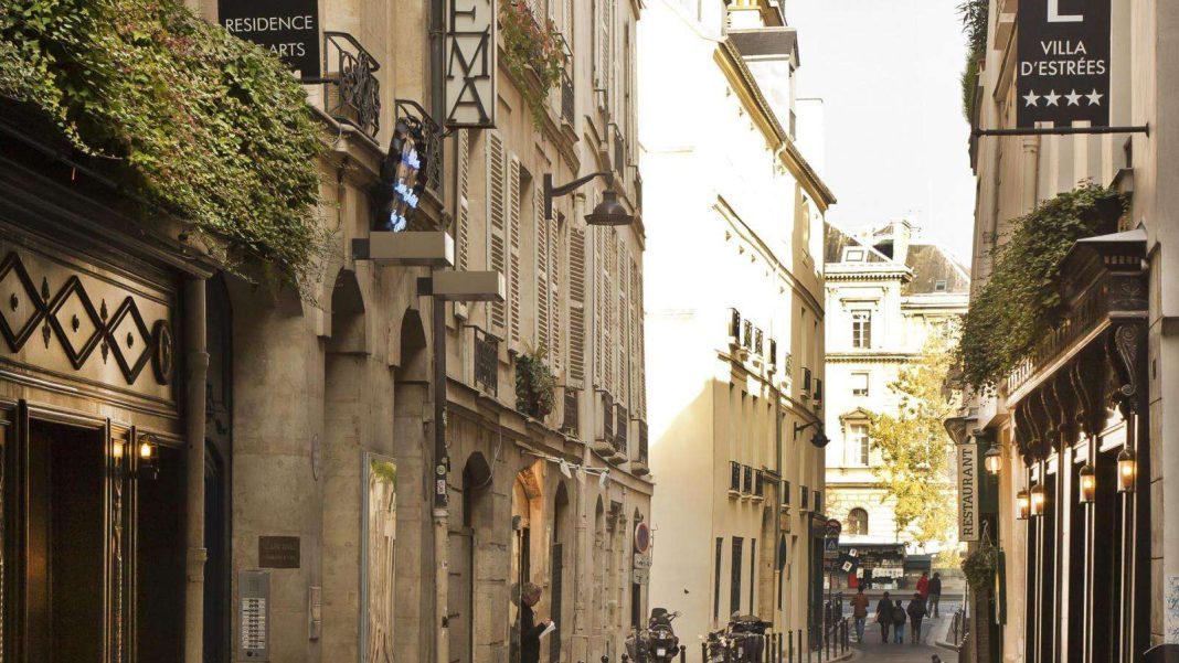 Une rue parisienne étroite bordée de bâtiments historiques ; des enseignes d'hôtels de charme comme la Résidence des Arts et l'Hôtel Villa d'Estrees sont suspendues au-dessus, avec de la verdure tombant en cascade des fenêtres. Des bicyclettes longent la rue pavée, éclairées par la lumière du soleil. 
