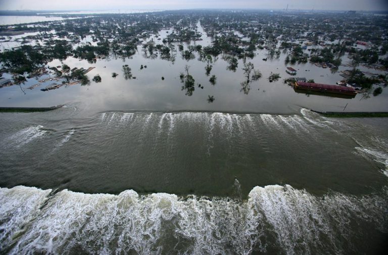 Vue aérienne d'une ville inondée, l'eau se déversant par-dessus une digue au premier plan. Des quartiers, des arbres et des bâtiments - dont certains sont connus pour leur architecture de restauration - sont submergés, et les eaux s'étendent au loin sous un ciel nuageux. 