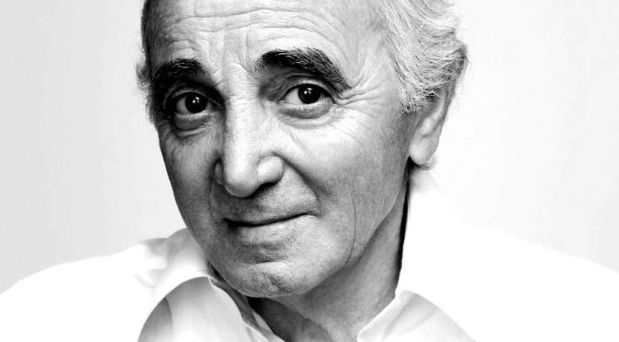 Charles Aznavour : « je veux que mon départ soit joliment orchestré » A black-and-white portrait against a soft, plain background of Charles Aznavour,as an older man with white hair and thick eyebrows, in a white collared shirt. He looks directly at the camera with a gentle, slight smile and laughter lines begining to crease his cheeks.