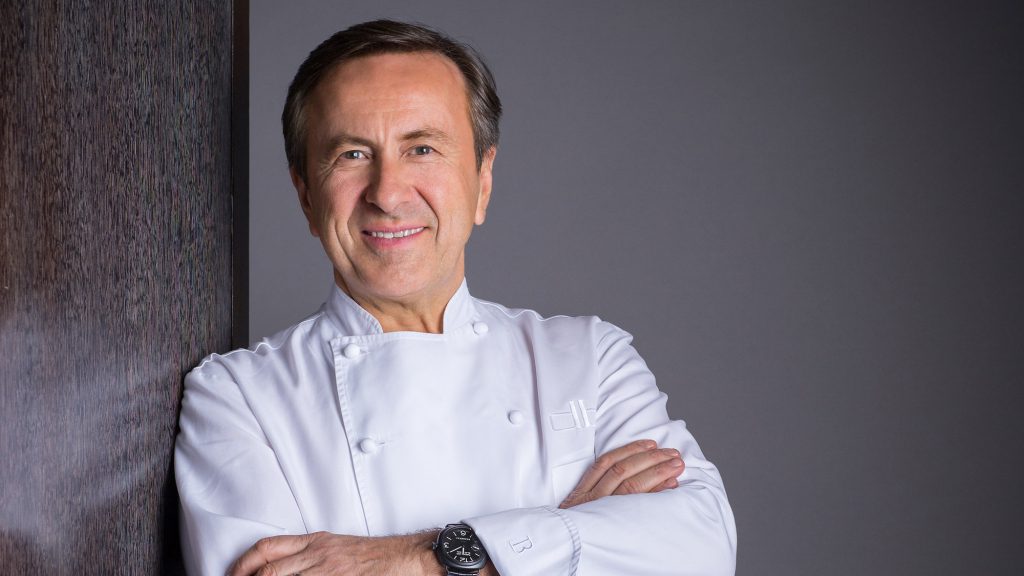 daniel-boulud-03 Un chef souriant d'âge moyen aux cheveux bruns se tient debout, les bras croisés, vêtu d'une blouse blanche et d'une montre noire. Il se tient contre un mur en bois sombre sur un fond gris uni, capturant un moment de la vie d'un chef.