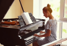 Qui n’a pas rêvé d’apprendre le piano ? A woman in a light blue lace dress sits at a grand piano, playing music. Sheet music is open on the stand as she continues to apprendre le piano, bathed in the warm, soft glow of sunlight streaming through large windows.