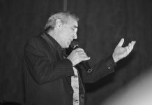 Aznavour : la biographie de référence Black and white photo of an older man with gray hair, in a dark pinstripe suit, singing passionately—his left hand raised, eyes closed. A true référence, he channels Aznavour’s spirit with every note. The background is out of focus.