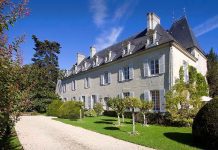 Château de Détilly The grand Château de Détilly boasts pale stone walls and tall, white-framed windows, nestled beside a gravel driveway. Manicured bushes and small trees adorn the lush green lawn beneath a clear sky with wispy clouds.