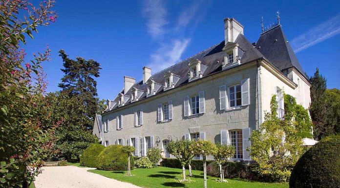 Château de Détilly The grand Château de Détilly boasts pale stone walls and tall, white-framed windows, nestled beside a gravel driveway. Manicured bushes and small trees adorn the lush green lawn beneath a clear sky with wispy clouds.