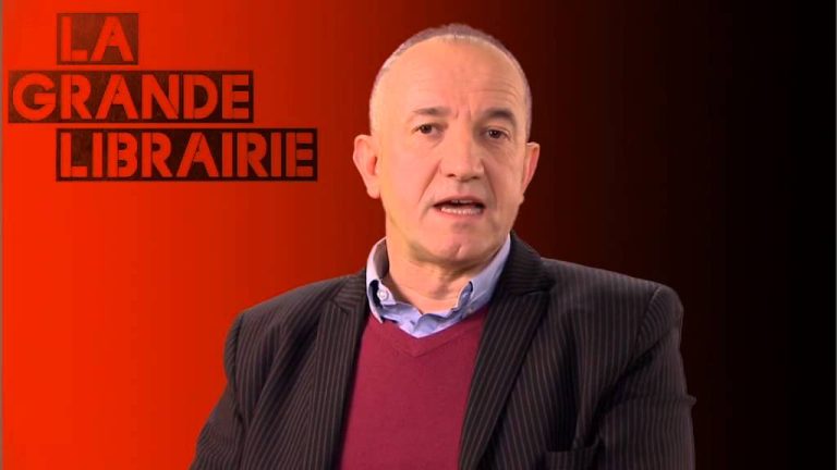 A middle-aged man in a striped dark suit jacket, light blue shirt, and burgundy sweater sits before a red and black gradient background. “LA GRANDE LIBRAIRIE” and “J'aime l'humain – Philippe Claudel” appear in bold red and black letters on the left.