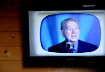 L’âge d’or de la chanson française A television mounted on a wooden wall displays an older man with gray hair, evoking the âge d'or of chanson française, speaking into a microphone. He wears a dark suit, red tie, and blue shirt against a blue background on a retro-styled TV screen.