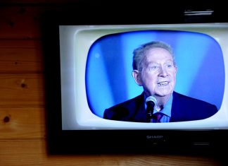 L’âge d’or de la chanson française A television mounted on a wooden wall displays an older man with gray hair, evoking the âge d'or of chanson française, speaking into a microphone. He wears a dark suit, red tie, and blue shirt against a blue background on a retro-styled TV screen.