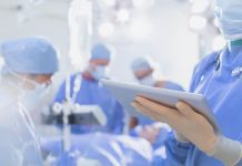 Traducteur médical Four surgeons in blue surgical scrubs and masks operate in a bright, sterile room. One surgeon holds a tablet, possibly using a traducteur médical app, highlighting teamwork and technology in the modern medical environment.