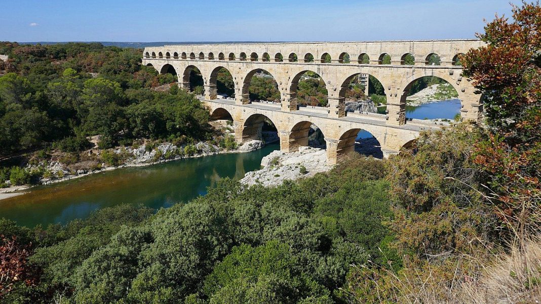 pont-du-gard Ancient Roman stone aqueduct bridge with multiple arches spans a river, surrounded by green trees and bushes under a clear blue sky. The bridges reflection is visible in the calm water below.