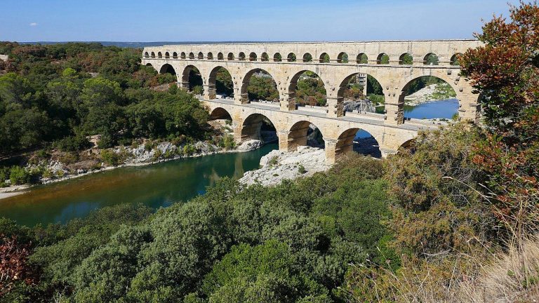 Un ancien pont aqueduc romain en pierre aux multiples arches enjambe une rivière, entouré d'arbres et de buissons verts sous un ciel bleu clair. Le reflet du pont est visible dans l'eau calme en contrebas. 