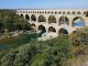 OCCITANIE Ancient Roman stone aqueduct bridge with multiple arches spans a river, surrounded by green trees and bushes under a clear blue sky. The bridges reflection is visible in the calm water below.
