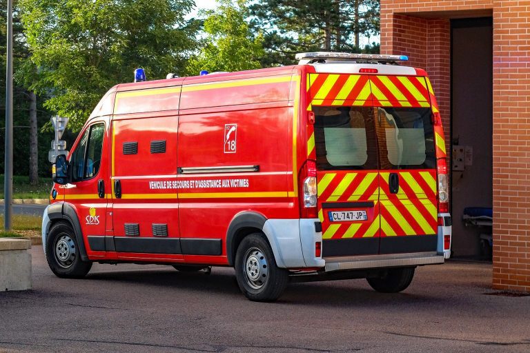 Un véhicule de secours d'urgence rouge vif, faisant partie des services d'urgence français, avec des chevrons jaunes et rouges, est garé devant un bâtiment en briques. L'inscription "Véhicule de Secours et d'Assistance aux Victimes" figure sur le côté. Des arbres verts se dressent sous un ciel ensoleillé. 