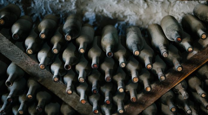How to manage a wine cellar Rows of dusty wine bottles are stored horizontally on wooden racks against a rough, white stone wall, highlighting expert wine cellar management. The aged bottles emphasize tradition and careful wine storage in this time-honored setting.