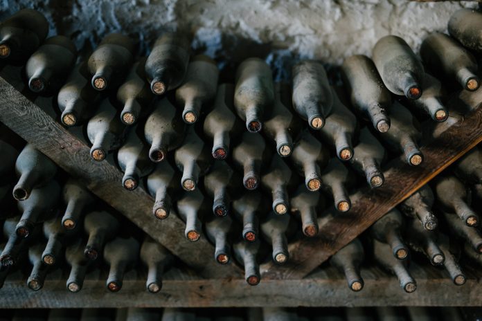 Rows of dusty wine bottles are stored horizontally on wooden racks against a rough, white stone wall, highlighting expert wine cellar management. The aged bottles emphasize tradition and careful wine storage in this time-honored setting.