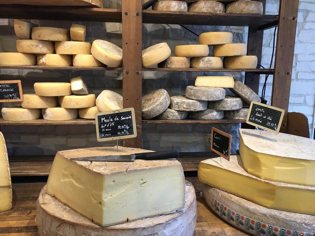 cheese Display Shelves stacked with various rounds and wheels of French cheese, some labeled by cheese families. On the wooden counter are large cut wedges, each marked with small black signs. A cheese knife rests on one wedge in this inviting cheese shop.