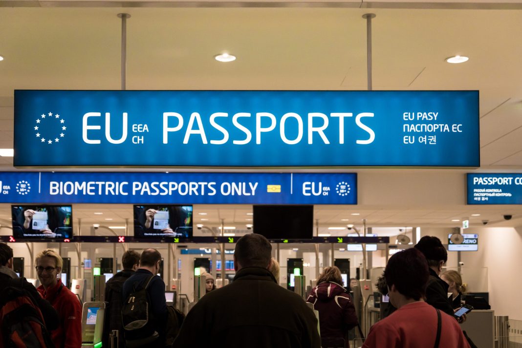People stand in line at an airport passport control area under a bright blue “EU EEA CH PASSPORTS” sign, with smaller text for translations and “BIOMETRIC PASSPORTS ONLY.” Automated scanners process travelers entering the Schengen Zone.