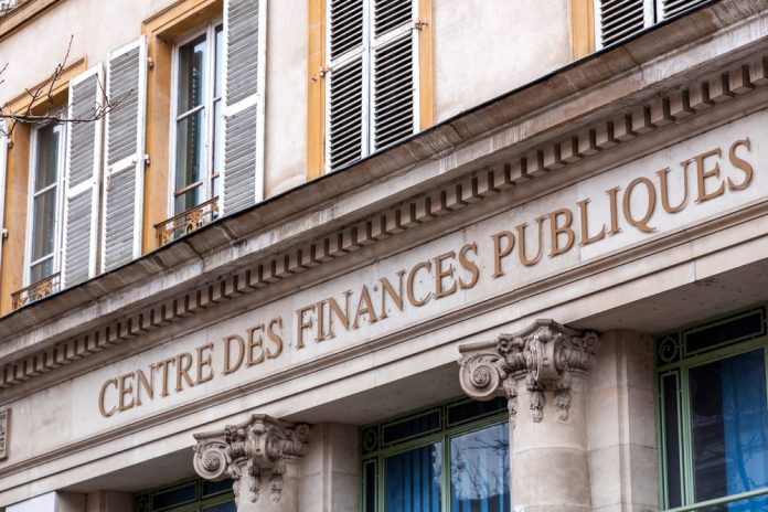 A stone building facade with large letters reading “CENTRE DES FINANCES PUBLIQUES” signifies where French property owners fulfill their mandatory declaration, featuring ornate columns and windows with open white shutters above. The architecture is classical and viewed from below.