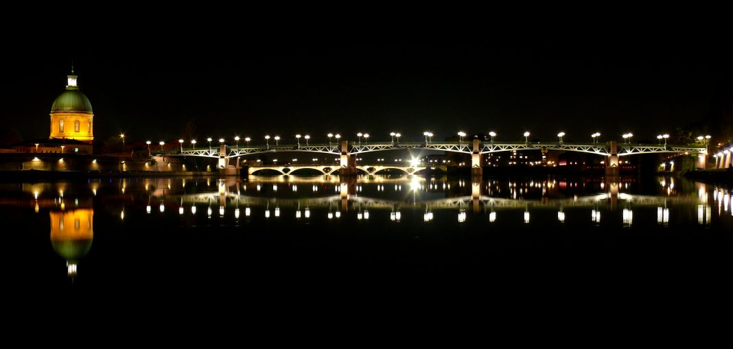 Le pont Saint-Pierre illuminé enjambant la Garonne au crépuscule, avec le dôme en cuivre emblématique de l'Hôpital de La Grave se reflétant dans l'eau.