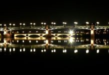 Visitors Guide to the City of Toulouse The illuminated Pont Saint-Pierre bridge spanning the Garonne River at twilight with the iconic copper dome of the Hôpital de La Grave reflecting in the water.