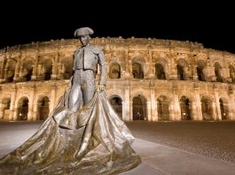 Guide Touristique de Nîmes Roman amphitheatre in Nimes brightly illuminated against a dark black starless night sky.In the foreground is a statue of a bullfighter holding his cape so it hangs below his knees and spreads out on the ground.The photo is taken at the foot of the bullfighter making him look taller than the amphitheatre in the background.