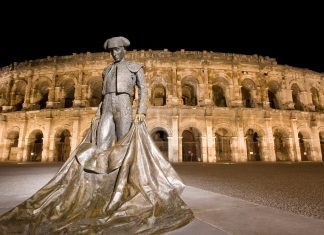 Guide Touristique de Nîmes Roman amphitheatre in Nimes brightly illuminated against a dark black starless night sky.In the foreground is a statue of a bullfighter holding his cape so it hangs below his knees and spreads out on the ground.The photo is taken at the foot of the bullfighter making him look taller than the amphitheatre in the background.