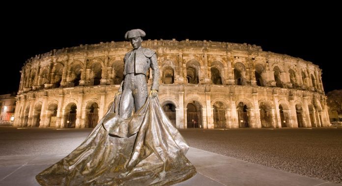 Roman amphitheatre in Nimes brightly illuminated against a dark black starless night sky.In the foreground is a statue of a bullfighter holding his cape so it hangs below his knees and spreads out on the ground.The photo is taken at the foot of the bullfighter making him look taller than the amphitheatre in the background.